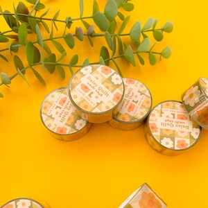 May include: A collection of five rolls of washi tape with a floral pattern. The tape is yellow, orange, and white with a daisy design. The text on the tape reads "Daisy Quilt washi tape Laurel mae art".