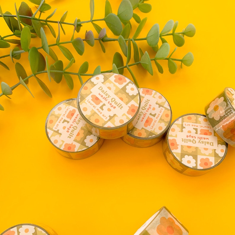 May include: A collection of five rolls of washi tape with a floral pattern. The tape is yellow, orange, and white with a daisy design. The text on the tape reads "Daisy Quilt washi tape Laurel mae art".