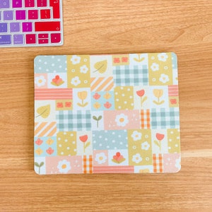 May include: A rectangular mouse pad featuring a patchwork design of various pastel-colored squares. The pattern includes light blue, yellow, and peach sections with motifs of white flowers, green leaves, orange tulips, and checkered or polka dot textures.
