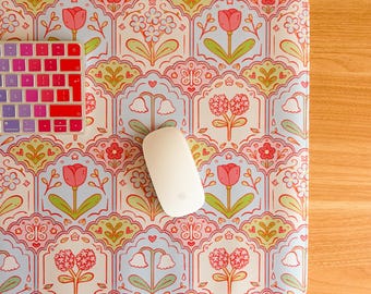 May include: A rectangular desk mat featuring a repeating pattern of pink tulips, blue flowers, and green leaves inside scalloped arches on a light blue background. A white computer mouse and a multi-colored keyboard are placed on the mat.