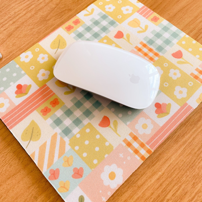 May include: A smooth white computer mouse rests on a rectangular mouse pad featuring a patchwork design of yellow, orange, and light green squares. The pattern includes illustrations of small white flowers, orange tulips, green leaves, and gingham checks.