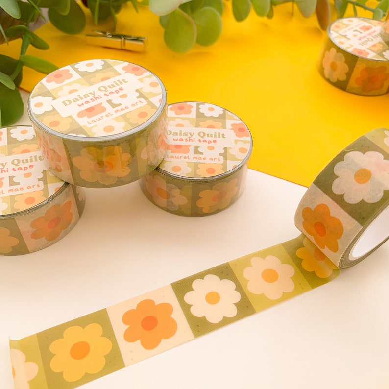 May include: Four rolls of washi tape with a floral pattern. The tape is green, yellow, and white with a daisy design. The tape is labeled "Daisy Quilt washi tape Laurel mae art".