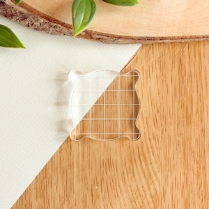 May include: A clear acrylic stamping block with a square shape and rounded corners, featuring a white grid pattern etched onto the surface to assist with alignment and positioning during the crafting process for stamps.