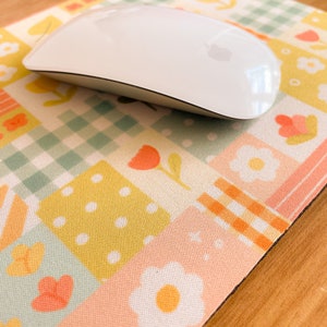 May include: A white computer mouse resting on a rectangular mouse pad featuring a patchwork design of pastel squares. The pattern includes light green gingham, yellow polka dots, and orange floral motifs on a soft, multi-colored grid background.