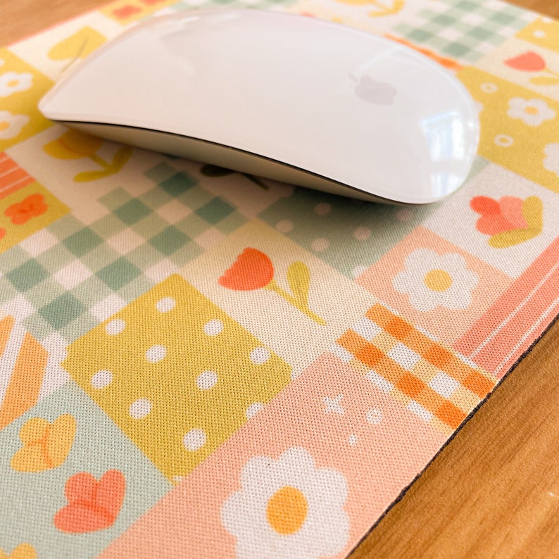 May include: A white computer mouse resting on a rectangular mouse pad featuring a patchwork design of pastel squares. The pattern includes light green gingham, yellow polka dots, and orange floral motifs on a soft, multi-colored grid background.