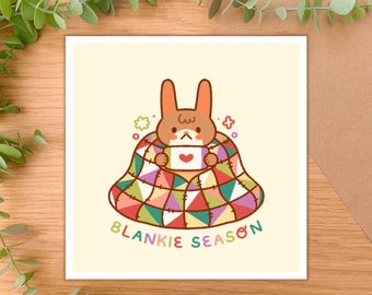 May include: A brown bunny wearing a colorful patchwork blanket, holding a cup with a heart on it. The text "BLANKIE SEASON" is below the bunny.