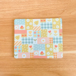 May include: A square mouse pad featuring a patchwork design of various pastel-colored rectangles. The pattern includes light blue, yellow, and pink sections decorated with white flowers, tulips, green leaves, polka dots, and blue or orange gingham checks.