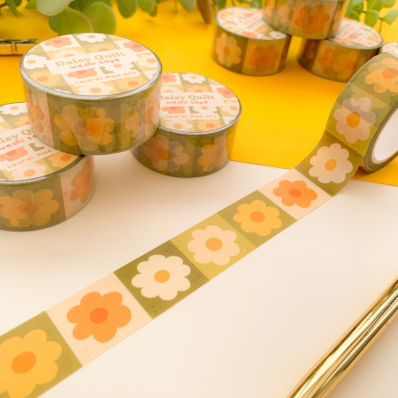 May include: A roll of green and yellow washi tape with a floral pattern. The tape has the text "Daisy Quilt washi tape Laurel mae art" printed on it.