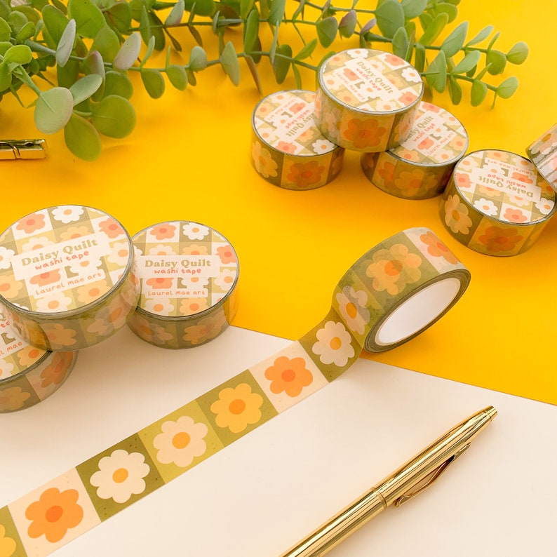 May include: A collection of floral patterned washi tape rolls with the text "Daisy Quilt washi tape Laurel mae art" printed on the rolls. The tape is green, yellow, and orange with a floral pattern.