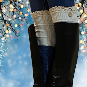 May include: A pair of black leather knee-high boots with buckles. The boots are worn with white knitted leg warmers with lace trim and a button.