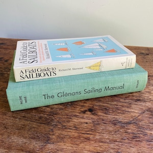 May include: Two hardcover books stacked on each other. The top book is A Field Guide to Sailboats by Richard M. Sherwood with a light blue cover featuring sailboat illustrations. The bottom book is The Glénans Sailing Manual in light green.