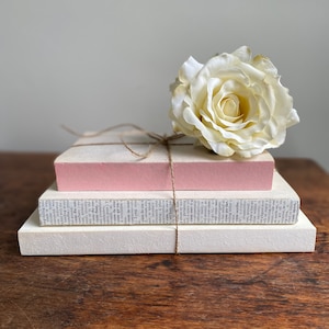 May include: A stack of three books with a pink book on top, a book with black text on the middle, and a white book on the bottom. A white rose is placed on top of the books. The books are tied together with twine.
