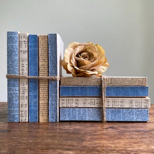 May include: A stack of five vertical books and a stack of four horizontal books wrapped in twine, featuring pages with printed text and musical notation in blue and beige tones, topped with a large, light brown artificial fabric rose.