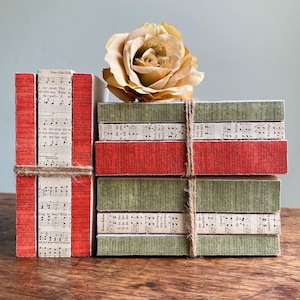 May include: Two stacks of books tied with twine. One stack is vertical with red spines and cream pages featuring musical notation. The other stack is horizontal with alternating olive green and cream spines, some with musical notation, topped by a beige rose.