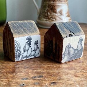 May include: Two wooden house-shaped blocks with rooster illustrations. The blocks have a rustic, weathered appearance with a dark wood grain. The rooster images are in black and white, adding a touch of farmhouse charm. The blocks are on a wooden surface.