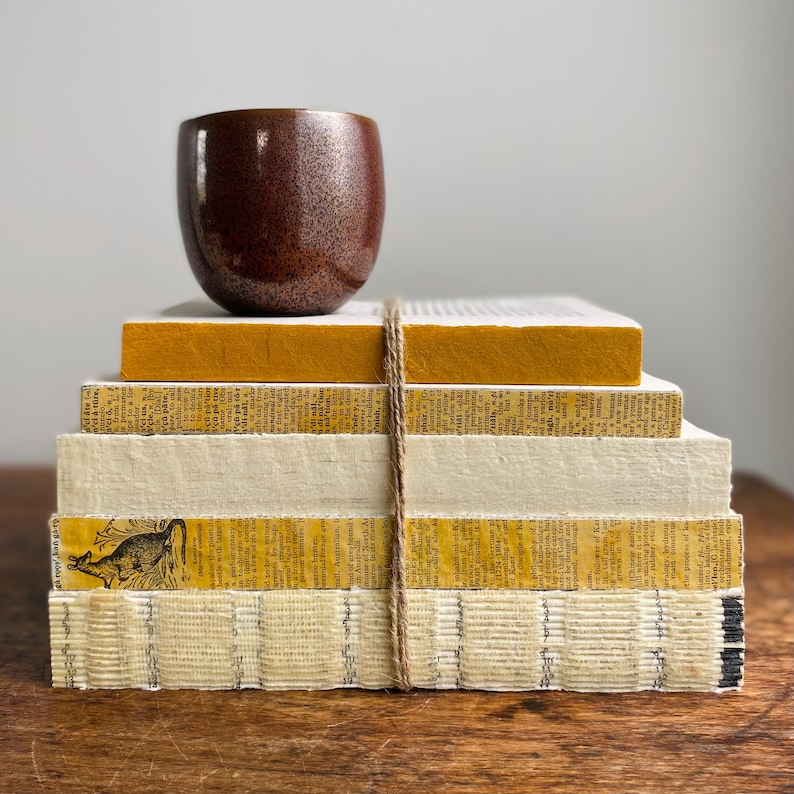 May include: A stack of vintage books with yellow and white covers, tied together with twine. A brown ceramic cup sits on top of the stack.