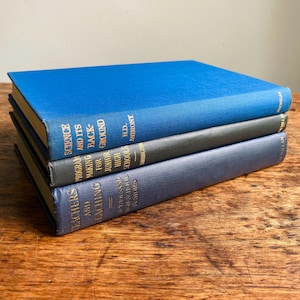 May include: A stack of three hardcover books with blue and grey cloth covers. The top book is bright blue, the middle is dark grey with gold text, and the bottom is navy blue with gold text. The spines show titles and author names clearly.