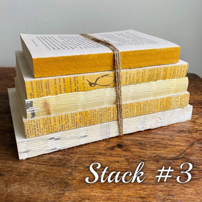 May include: A stack of five books with yellow and white covers, tied together with twine. The top book has a yellow spine and the rest have white spines. The books are on a wooden surface. Stack #3