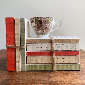 May include: A stack of decorative books tied with twine, topped with a vintage teacup. The books have red, green, and sheet music-covered spines. The teacup features a brown and white pastoral scene.