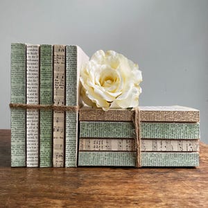 May include: A decorative set of bookends and a small stack of decorative boxes, all covered in vintage book pages and sheet music. A large, cream-colored rose sits atop the boxes. The items are tied with twine and rest on a wooden surface.