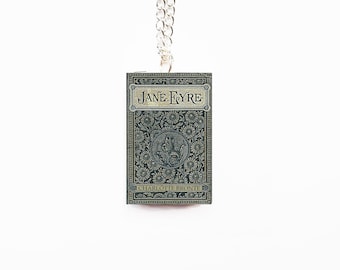 May include: A rectangular pendant necklace featuring a miniature book cover of "Jane Eyre" by Charlotte Brontë. The book cover is dark teal with floral and scroll designs, and the title is in a banner at the top. The pendant hangs from a silver chain.