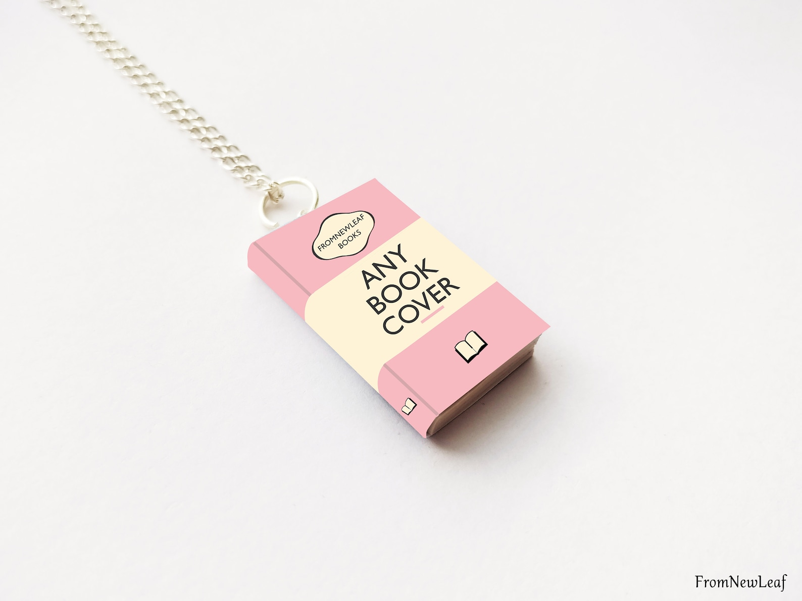 The pink charm-sized book with black text 'Any Book Cover' dangles from a silver chain with a metal ring attached.