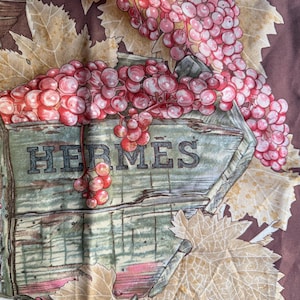 May include: A silk scarf with a detailed illustration of a wooden cart overflowing with ripe, pink grapes. The word "HERMES" is printed on the cart. The design includes tan-coloured leaves against a dark brown background, creating a vintage aesthetic.