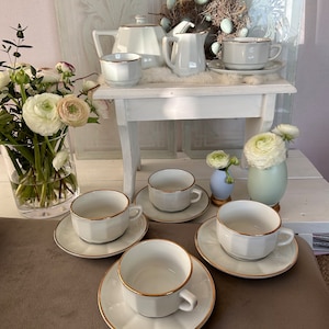 May include: A white tea set with gold trim, including a teapot, creamer, sugar bowl, and four teacups and saucers. The tea set is arranged on a white table with a floral arrangement in the background.