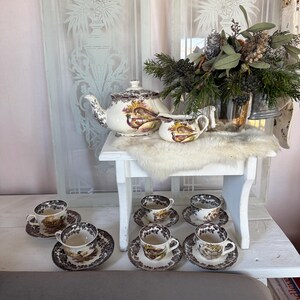 Op de afbeelding: Een vintage theeservies met een theepot, melkkan, suikerpot en zes theekopjes met schotels. De set heeft een bruin en wit vogel- en bloemmotief. Het theeservies staat op een witte tafel met een imitatiebont.