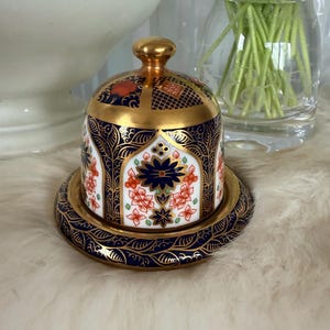 Puede incluir: Una mantequera de cerámica decorativa con tapa, con un diseño azul marino y dorado. El plato tiene un patrón floral con detalles en rojo y blanco. La tapa está rematada con un pomo dorado y descansa sobre un platillo a juego.