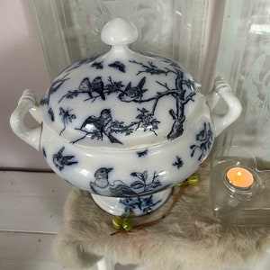 Puede incluir: Fuente de porcelana blanca con tapa, con un diseño de pájaros y ramas en azul y blanco. La fuente tiene dos asas y un pomo en forma de hongo en la tapa. Una pequeña vela en un portavelas de cristal está al lado.