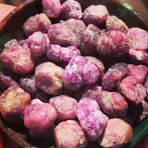 Op de afbeelding: Een kom gevuld met verschillende roze en paarse edelstenen. De stenen zijn ruw en hebben een natuurlijke, onbewerkte uitstraling.