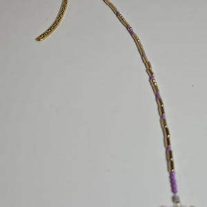 May include: A gold-colored metal bookmark with a decorative swirl design. The bookmark has a beaded charm with gold and purple beads, and a pink butterfly charm at the end. The bookmark is designed for books.