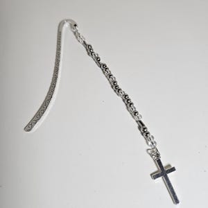 May include: A silver bookmark with a decorative curved top and a beaded chain. The chain is composed of small silver and clear beads, leading to a silver cross pendant. The bookmark lies on a white surface.