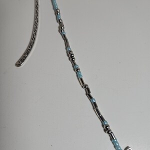 May include: A silver metal bookmark with a decorative curved design on the top portion. The bottom features a beaded strand with silver and light blue beads, and a silver butterfly charm.
