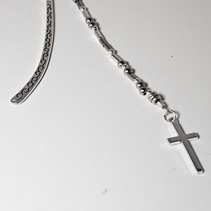 May include: A silver metal bookmark with a cross charm. The bookmark has a curved top and a decorative design along the edge. The charm is attached to a beaded chain.