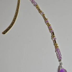 May include: A gold-colored metal bookmark with a decorative curved design. The bookmark has a beaded strand of gold and purple beads, ending with a purple teardrop-shaped charm. The beads are strung on a silver-colored wire.