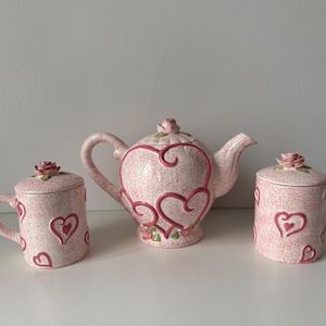 May include: A pink and white ceramic tea set with a teapot and two mugs. The teapot and mugs are decorated with pink hearts and roses.