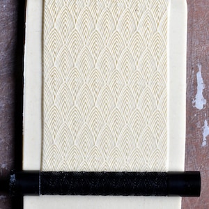 May include: A white fondant sheet with a raised geometric pattern of overlapping arches. A black rolling pin is placed on top of the fondant.