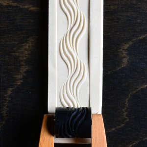 May include: A white rolling pin with a black rubber roller with a wavy pattern. The roller is attached to a wooden handle.