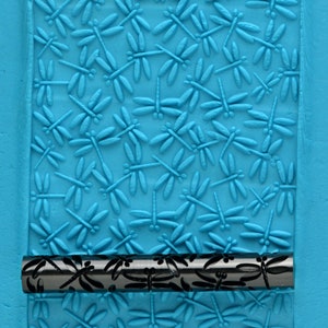 May include: A black rolling pin with a dragonfly design. The rolling pin is used to create an embossed pattern on fondant or clay.