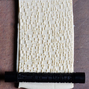 May include: A white rolling pin with a textured pattern, used for creating decorative designs in dough. The rolling pin is lying on a wooden surface and a black rolling pin handle is attached to the end.