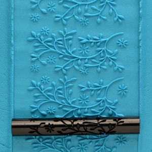 May include: A black rolling pin with a floral design on a blue background. The rolling pin is used for baking and decorating.