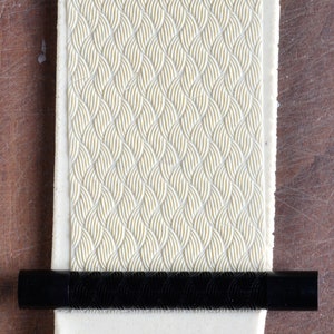 May include: A white rolling pin with a raised pattern of woven lines. The rolling pin is on a wooden surface and a black rolling pin is in front of it.