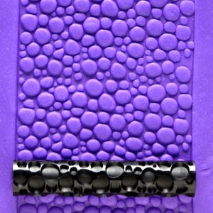 May include: A close-up of a purple silicone rolling pin with a textured pattern of small circles. The rolling pin is designed to create a pebble-like texture on fondant or other edible materials.
