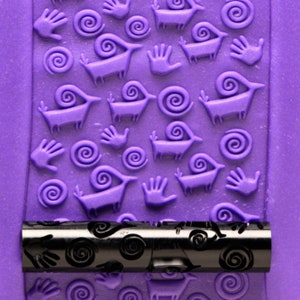 Peut inclure: Un gros plan d'un rouleau à pâtisserie violet avec un motif répété de mains, de spirales et de formes d'animaux stylisées. Le rouleau à pâtisserie est en métal et a une poignée noire.