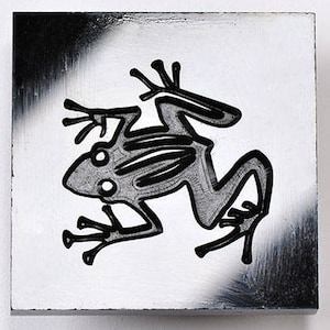 May include: Square, white painted wooden block with a stylized black and gray frog illustration. The frog is outlined in black, with a gray interior. The background has a white base with black paint strokes in the corners.