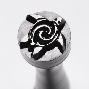 May include: Close-up of a metal stamp with a detailed turtle design. The turtle is depicted in a stylized, swirling pattern, with a dark, contrasting background. The stamp is circular and mounted on a cylindrical handle.