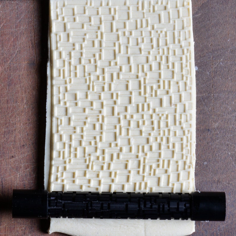May include: A white rolling pin with a textured pattern, used for creating decorative designs in dough. The rolling pin is lying on a wooden surface and a black rolling pin handle is attached to the end.
