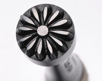 May include: A black metal stamp with a daisy design. The stamp has a round head and a cylindrical handle.